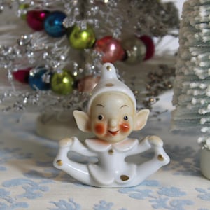 May include: A vintage ceramic Christmas elf figurine in a seated pose, wearing a white outfit with gold accents and rosy cheeks. The elf is set against a backdrop of a silver tinsel Christmas tree with colorful ornaments.