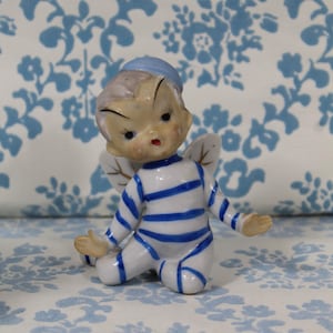 May include: A vintage ceramic figurine of a seated cherub with blue and white striped clothing and a matching cap. The cherub has small, delicate wings and is set against a floral patterned background.