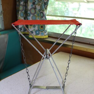 May include: A retro folding camp stool featuring a red seat and a silver metal frame. The stool has a triangular design and is supported by chains. The background includes a window, curtains, and a table.
