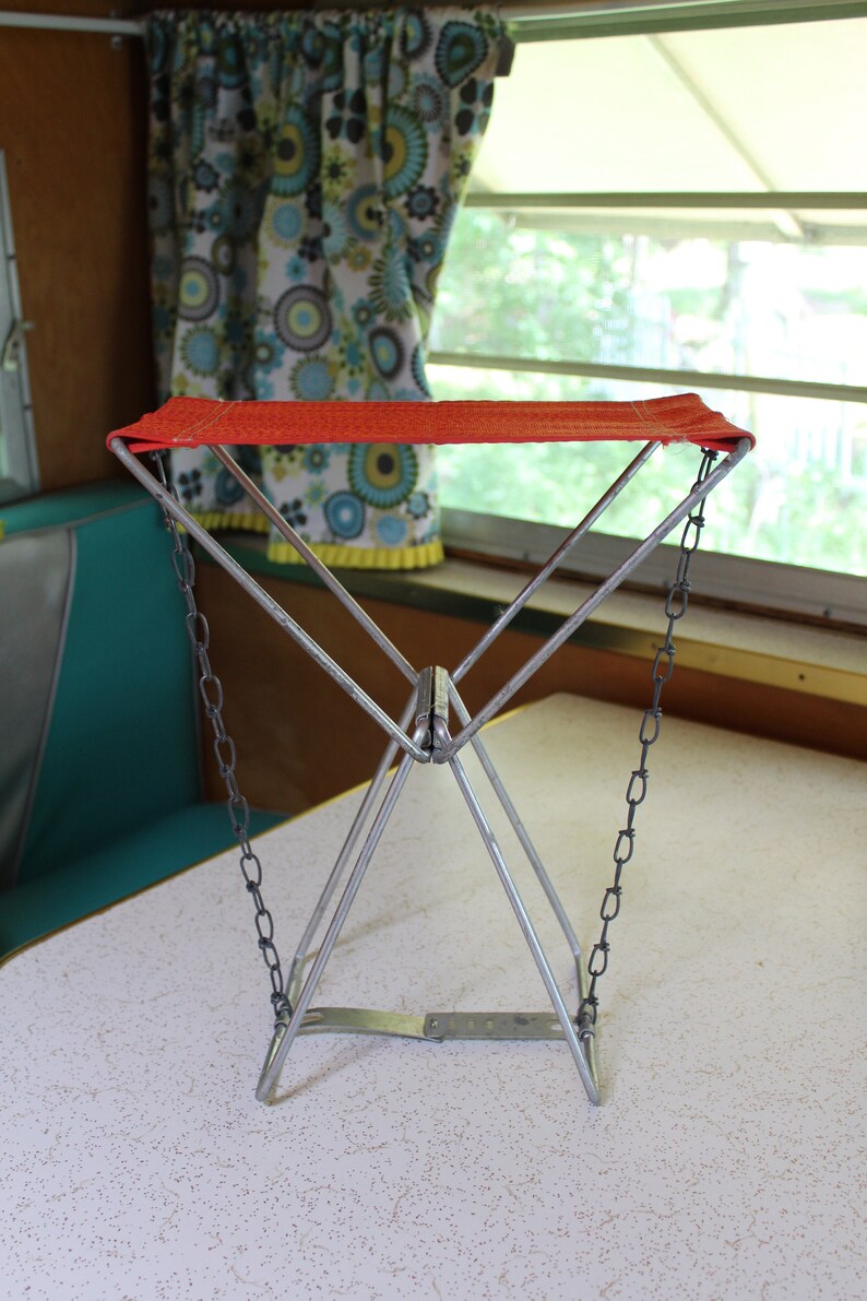 May include: A retro folding camp stool featuring a red seat and a silver metal frame. The stool has a triangular design and is supported by chains. The background includes a window, curtains, and a table.