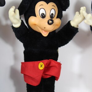 May include: Vintage Mickey Mouse plush doll with a black body, red shorts, and brown shoes. The doll has a tan face, white gloves, and black ears. A yellow button with a black "X" is on the shorts.