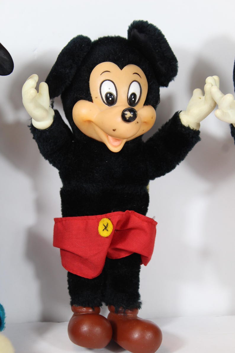 May include: Vintage Mickey Mouse plush doll with a black body, red shorts, and brown shoes. The doll has a tan face, white gloves, and black ears. A yellow button with a black "X" is on the shorts.
