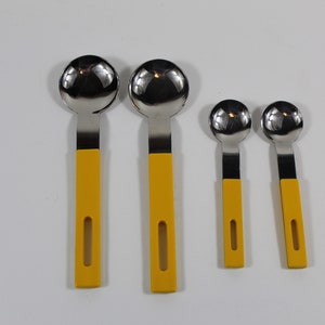 May include: Four stainless steel measuring spoons with yellow handles. The spoons are arranged in order of size, with the largest spoon on the left and the smallest spoon on the right.