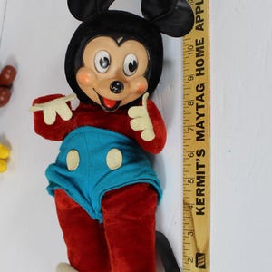 May include: Vintage Mickey Mouse plush doll with a black head and ears, red body, blue shorts, and white gloves and shoes. The doll is standing next to a wooden ruler.