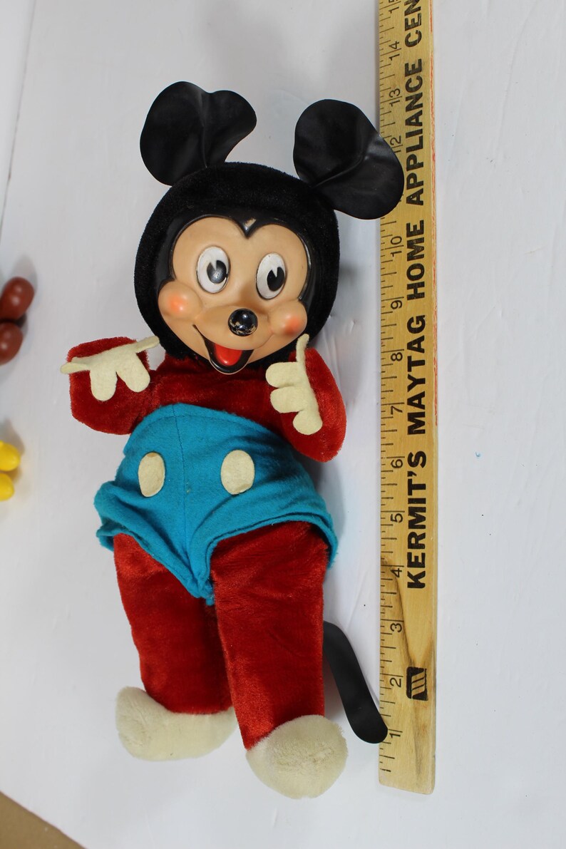 May include: Vintage Mickey Mouse plush doll with a black head and ears, red body, blue shorts, and white gloves and shoes. The doll is standing next to a wooden ruler.