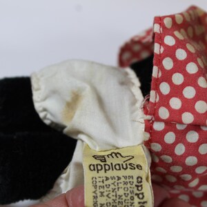 May include: Close-up of a vintage plush toy with black fur, yellow feet, and a red polka dot bow. A white ruffled collar and a yellow tag with the word "applause" are also visible.