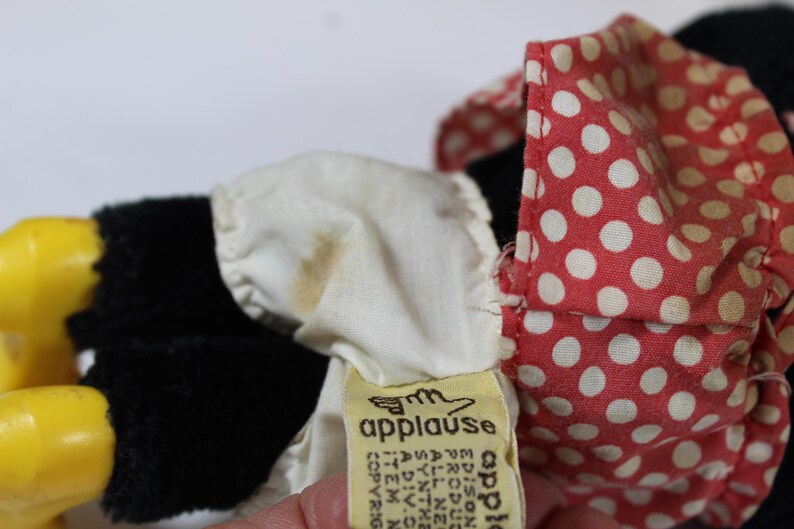 May include: Close-up of a vintage plush toy with black fur, yellow feet, and a red polka dot bow. A white ruffled collar and a yellow tag with the word "applause" are also visible.