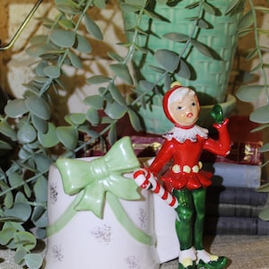 May include: A white ceramic Christmas mug with a green bow and a candy cane. A red and green elf figurine is sitting on the mug.