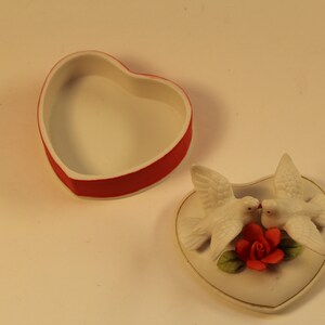 May include: A heart-shaped trinket box with a red rim and white interior sits open next to a heart-shaped lid. The lid features two white doves kissing above a red rose.