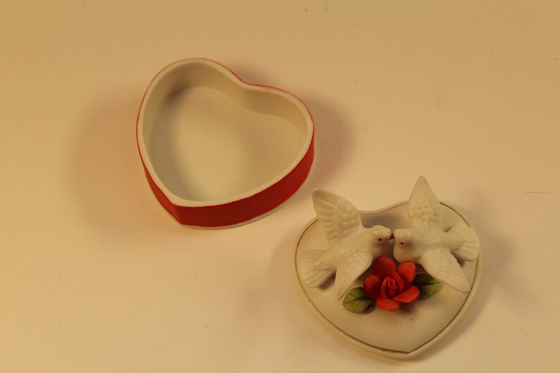 May include: A heart-shaped trinket box with a red rim and white interior sits open next to a heart-shaped lid. The lid features two white doves kissing above a red rose.