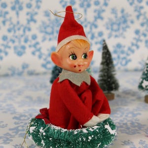 May include: A vintage Christmas elf figurine. The elf is dressed in a red felt suit and hat with a white trim. It has a painted face with green eyes and is seated on a green wreath with faux snow. Miniature Christmas trees are in the background.