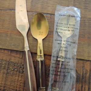 May include: A set of vintage bronze spoons and a butter knife with dark wood handles and brass accents. One spoon is in a plastic sleeve with text. The set is on a wooden surface.