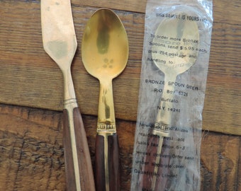 May include: A set of vintage bronze spoons and a butter knife with dark wood handles and brass accents. One spoon is in a plastic sleeve with text. The set is on a wooden surface.