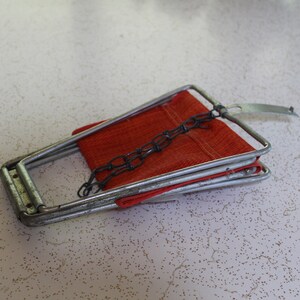 May include: A vintage metal folding chair with a red fabric seat. The chair has a silver frame and a chain detail. The chair is folded and resting on a speckled white surface.