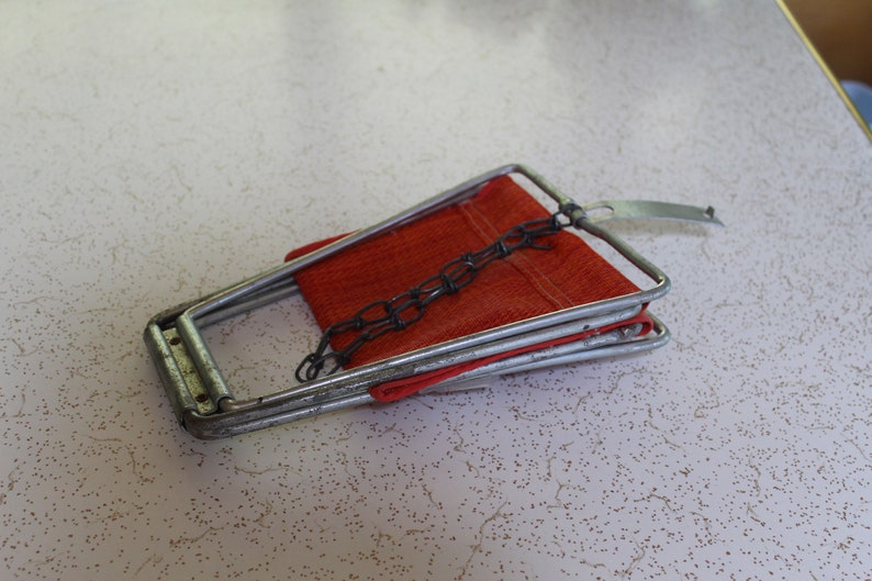 May include: A vintage metal folding chair with a red fabric seat. The chair has a silver frame and a chain detail. The chair is folded and resting on a speckled white surface.