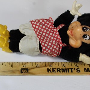May include: A vintage Minnie Mouse plush doll, lying down, with a black body, white gloves, and yellow shoes. She wears a red polka dot dress and bow. A wooden ruler with the text "KERMIT'S MAYTAG" is in the foreground.