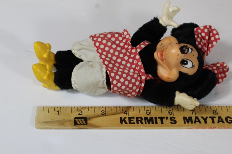 May include: A vintage Minnie Mouse plush doll, lying down, with a black body, white gloves, and yellow shoes. She wears a red polka dot dress and bow. A wooden ruler with the text "KERMIT'S MAYTAG" is in the foreground.