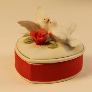 May include: A heart-shaped trinket box with a red base and white lid, adorned with two white doves and a red rose. The box has a gold trim and is likely made of ceramic or porcelain.