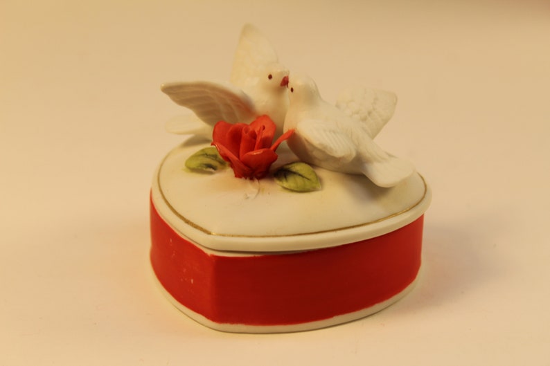 May include: A heart-shaped trinket box with a red base and white lid, adorned with two white doves and a red rose. The box has a gold trim and is likely made of ceramic or porcelain.
