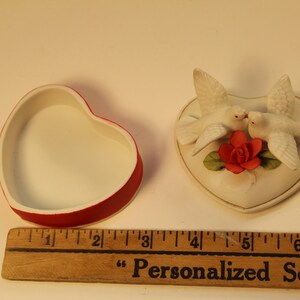 May include: A heart-shaped trinket box with a red rim and white interior sits next to a heart-shaped decorative piece. The decorative piece features two white doves kissing above a red rose. The box measures approximately 2 inches.