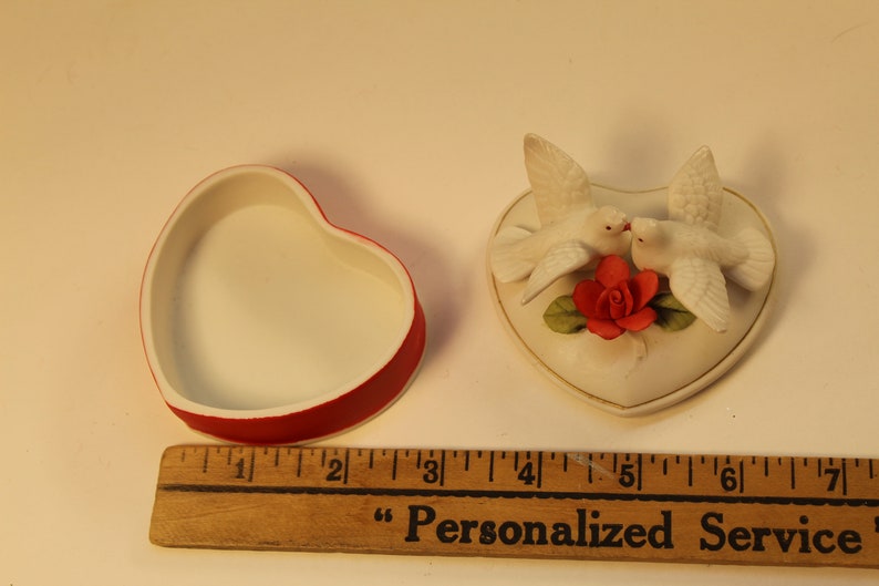 May include: A heart-shaped trinket box with a red rim and white interior sits next to a heart-shaped decorative piece. The decorative piece features two white doves kissing above a red rose. The box measures approximately 2 inches.