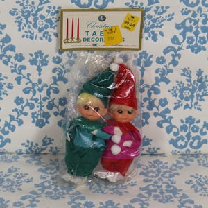 May include: A package of two Christmas table decorations. One elf is wearing a green outfit and hat, the other is wearing a red outfit and hat. The dolls have blonde hair and are packaged in clear plastic. The package has a yellow tag that reads "Christmas Table Decor".