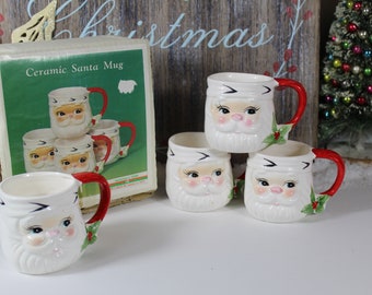 May include: Set of four white ceramic Santa mugs with red handles. Each mug features a Santa Claus face with a white beard and a red nose. The mugs are in their original box, which says "Ceramic Santa Mug".