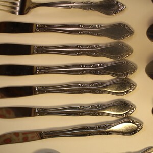 May include: A set of eight silver-toned flatware pieces, including a fork and seven knives. The knives have ornate handles with a decorative design.