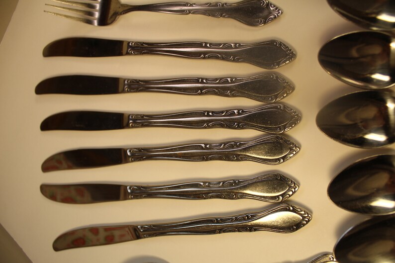 May include: A set of eight silver-toned flatware pieces, including a fork and seven knives. The knives have ornate handles with a decorative design.