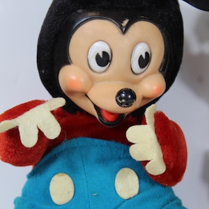 May include: Vintage Mickey Mouse plush doll. The doll features a black head and ears, a tan face with rosy cheeks, and a red and blue outfit. The doll's hands are white, and it has a cheerful expression.