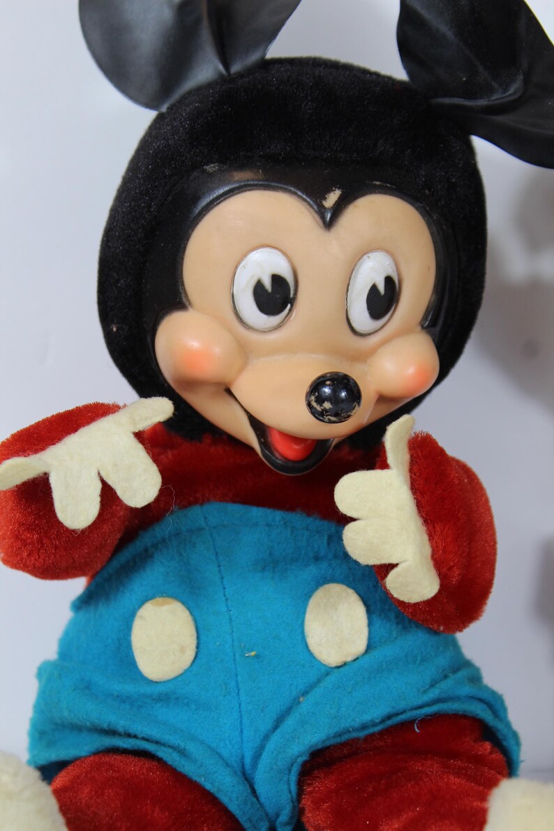 May include: Vintage Mickey Mouse plush doll. The doll features a black head and ears, a tan face with rosy cheeks, and a red and blue outfit. The doll's hands are white, and it has a cheerful expression.
