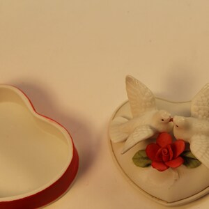 May include: A heart-shaped trinket box with a red rim and a white interior sits next to a heart-shaped decorative piece. The decorative piece features two white doves kissing above a red rose with green leaves.