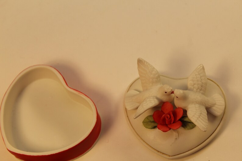 May include: A heart-shaped trinket box with a red rim and a white interior sits next to a heart-shaped decorative piece. The decorative piece features two white doves kissing above a red rose with green leaves.