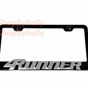 May include: A black license plate frame featuring the silver-colored text 4Runner embossed along the bottom edge. The frame includes two mounting holes at the top and displays the watermark text 3DShopByKelly in the upper left corner.