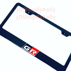 May include: A black rectangular license plate frame featuring a centered emblem with the letters GR in white on a red and black background. The frame includes two mounting holes at the top and displays the watermark text 3DShopByKelly.