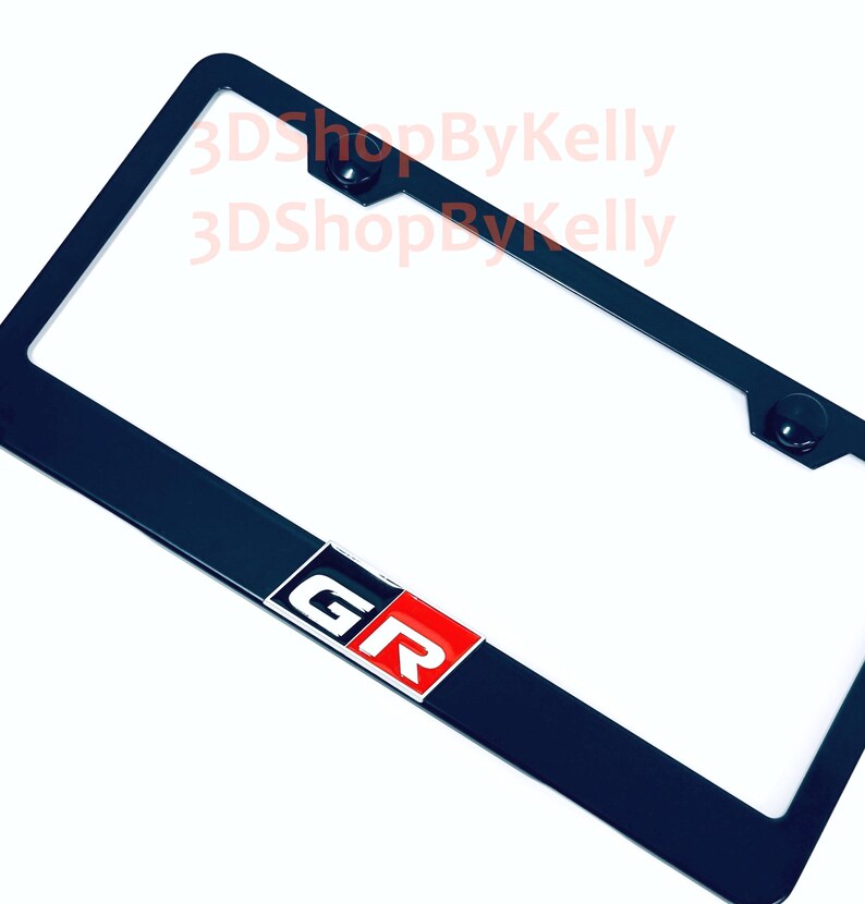 May include: A black rectangular license plate frame featuring a centered emblem with the letters GR in white on a red and black background. The frame includes two mounting holes at the top and displays the watermark text 3DShopByKelly.