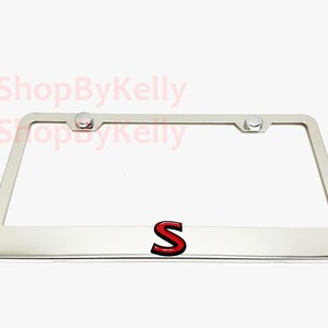 May include: A chrome license plate frame with a red and black letter "S" emblem. The frame has a rectangular shape with rounded corners and includes mounting hardware.