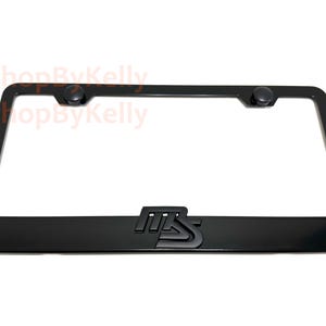 May include: A rectangular black license plate frame with rounded corners and two circular mounting caps at the top. The bottom center of the frame features a raised, stylized black emblem with the letters MS intertwined in a bold, blocky design.