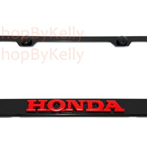 May include: Black license plate frame with the red "HONDA" logo. The frame has a rectangular shape with rounded corners and two black screw caps at the top.