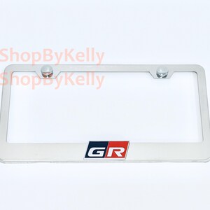 May include: A rectangular silver-colored metal license plate frame featuring the GR logo in black, white, and red at the bottom center. The frame includes two circular mounting holes at the top with silver hardware. The text 3DShopByKelly is visible.