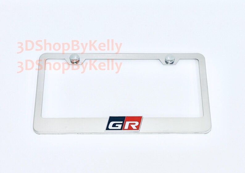 May include: A rectangular silver-colored metal license plate frame featuring the GR logo in black, white, and red at the bottom center. The frame includes two circular mounting holes at the top with silver hardware. The text 3DShopByKelly is visible.