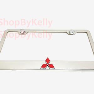 May include: A chrome license plate frame with a red and silver Mitsubishi logo. The frame has a rectangular shape with rounded corners and two mounting posts. The text "3DShopByKelly" is visible in the background.