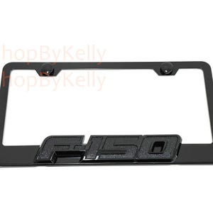 May include: Black license plate frame with the embossed letters "F-150". The frame has a rectangular shape with rounded corners and includes four mounting holes. The frame is made of a durable material and is designed to fit standard U.S. license plates.