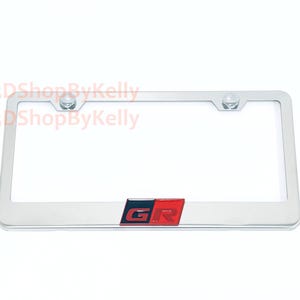 May include: A rectangular chrome license plate frame featuring a centered emblem with the letters GR in a bold font. The emblem is split into a black section on the left and a red section on the right. Two circular mounting holes are at the top.