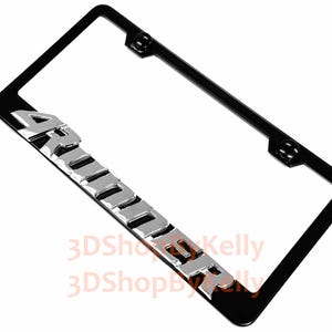 May include: A rectangular black license plate frame featuring the word ARMORER in raised, metallic silver-toned block lettering along the bottom edge. The frame includes two circular black mounting points at the top for secure vehicle attachment.