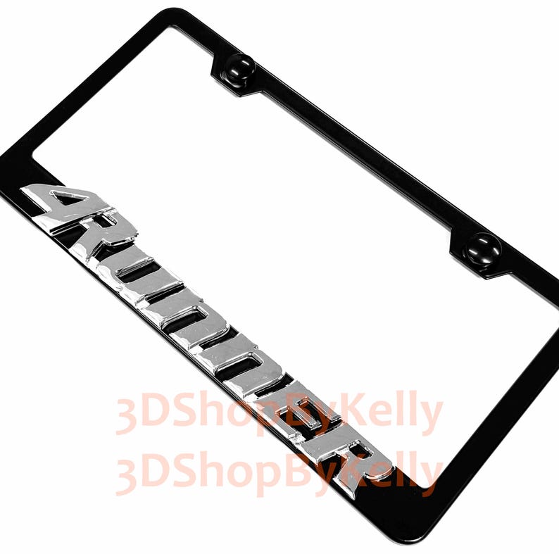 May include: A rectangular black license plate frame featuring the word ARMORER in raised, metallic silver-toned block lettering along the bottom edge. The frame includes two circular black mounting points at the top for secure vehicle attachment.