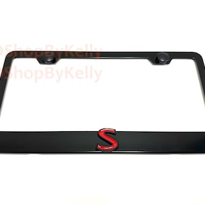 May include: Black license plate frame with a red and black "S" emblem. The frame has a rectangular shape with rounded corners and two black screw caps at the top. The background is white.