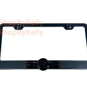 May include: A rectangular black license plate frame featuring a centered circular emblem with a stylized logo. The frame includes two circular mounting points at the top corners and a smooth, matte finish throughout the entire structure.