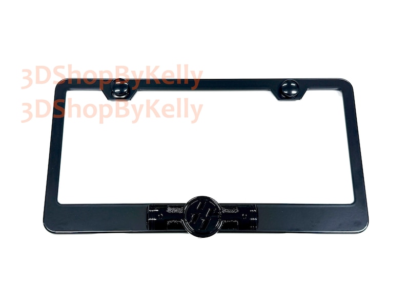 May include: A rectangular black license plate frame featuring a centered circular emblem with a stylized logo. The frame includes two circular mounting points at the top corners and a smooth, matte finish throughout the entire structure.