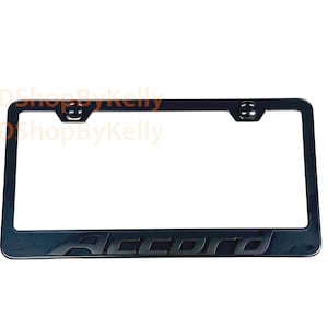 May include: A black rectangular license plate frame featuring the word Accord embossed in a matching black font along the bottom edge. The frame includes two circular mounting holes at the top with integrated black hardware caps for installation.